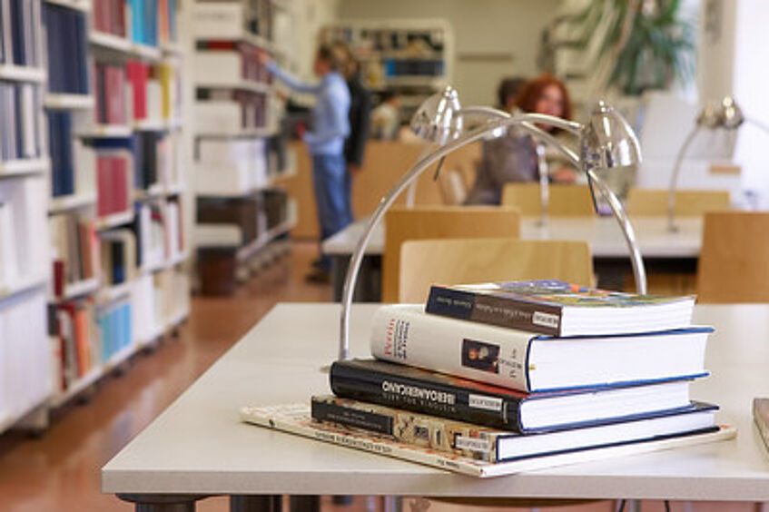 Ein Closeup eines Bücherstapels, im Hintergrund ist eine Bücherei zu sehen.