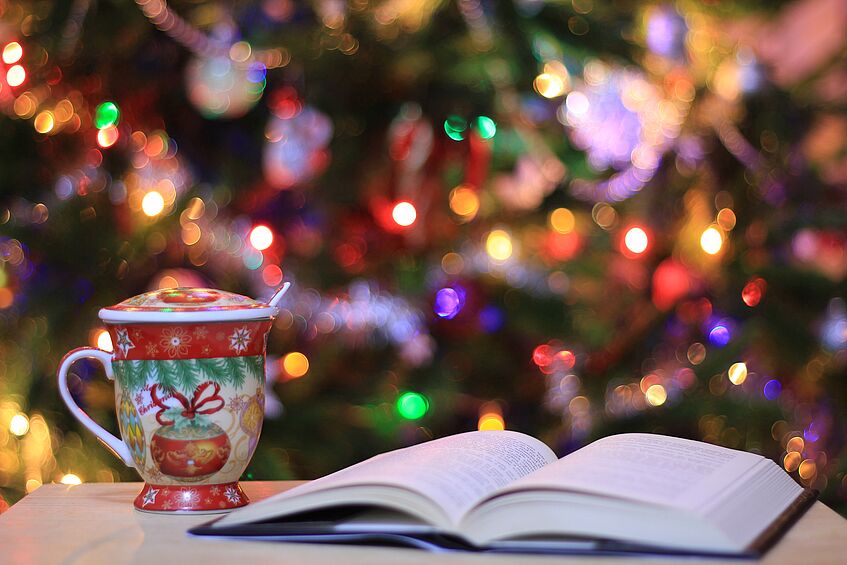 Ein aufgeschlagenes Buch und eine Tasse stehen auf einem Tisch vor einem bunt leuchtenden Weihnachtsbaum.