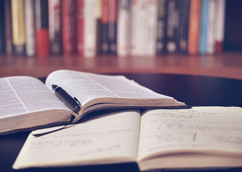 Ein offenes Buch sowie ein Notizbuch liegen auf einem Schreibtisch. Im Hintergrund sieht man ein Bücherregal.